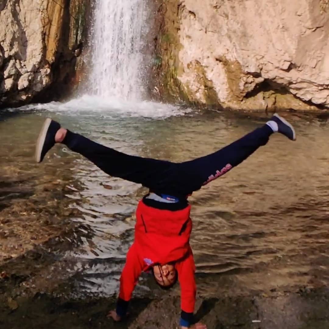 A student doing a wide-legged handstand by a waterfall.